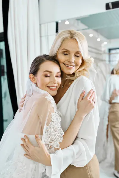 Une jeune mariée brune dans une robe de mariée et sa mère d'âge moyen s'embrassent devant un miroir dans un salon de mariée. — Photo de stock