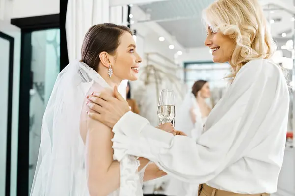 Une jeune mariée dans une robe de mariée blanche et sa mère, toutes deux élégamment vêtues de blanc, trinquent avec des verres de champagne. — Photo de stock