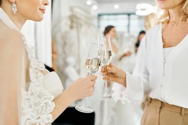 Une jeune mariée brune dans une robe de mariée se tient avec sa mère d'âge moyen, toutes deux tenant des verres de champagne dans un salon de mariée. — Photo de stock