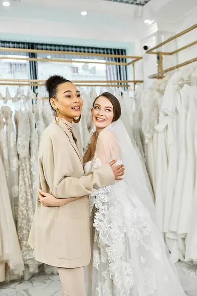 Uma jovem noiva morena e sua dama de honra ficam lado a lado em um salão de noivas, olhando para vários vestidos de noiva.. — Fotografia de Stock