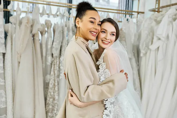 A young bride in a white wedding dress embracing her bridesmaid in front of a rack of various wedding dresses in a bridal salon. — Stock Photo