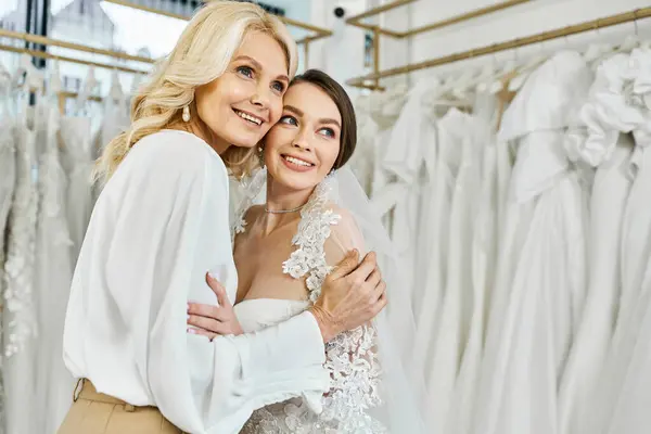 Eine junge brünette Braut im Brautkleid umarmt ihre Mutter mittleren Alters in einem Brautsalon voller Brautkleider. — Stockfoto
