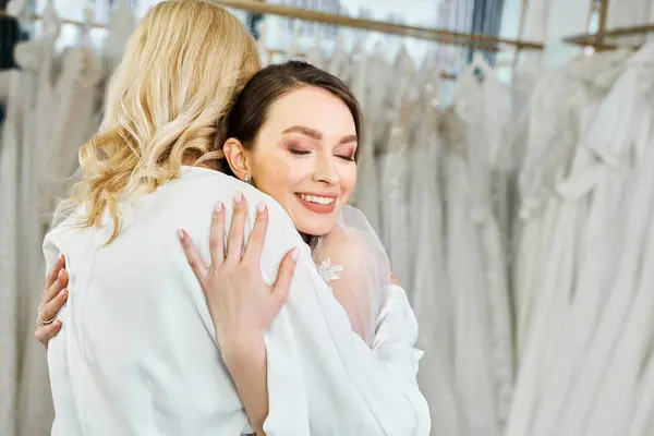 Una joven novia en un vestido de novia abraza a su madre de mediana edad en un salón de novias delante de un estante de vestidos. - foto de stock