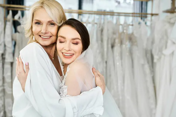 Две женщины, молодая брюнетка невеста в свадебном платье и ее мать средних лет, обнимаются перед стойкой платьев в свадебном салоне. — стоковое фото
