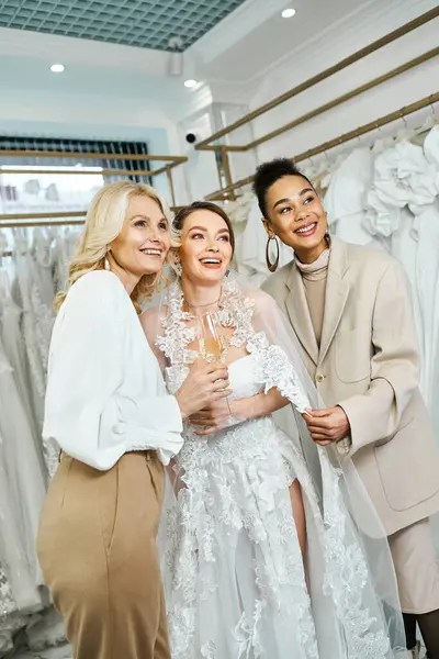 Eine junge Braut im Brautkleid, ihre Mutter mittleren Alters und ihre beste Freundin als Brautjungfer vor einem Kleiderständer. — Stockfoto