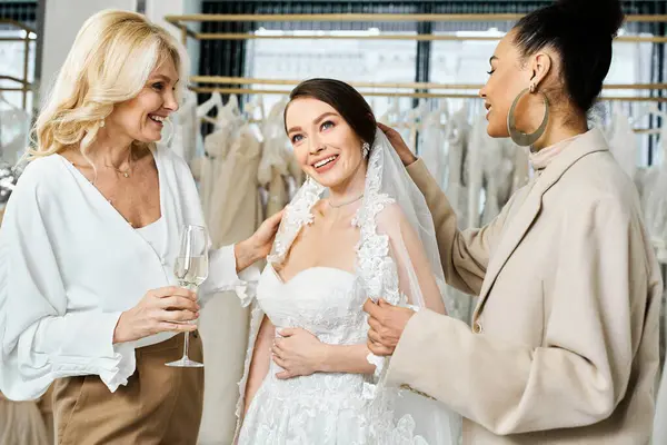 Жінки, наречена в білій весільній сукні, її мати, і її найкращий друг як подруга нареченої, стоять разом перед стійкою барвистих суконь. — стокове фото