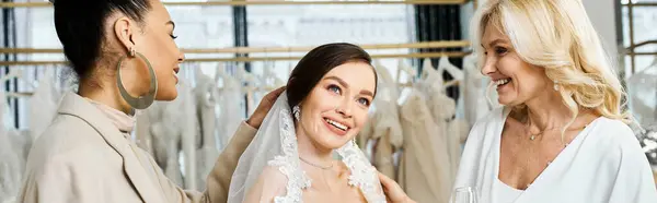 Uma jovem noiva morena, sua mãe de meia-idade e melhor amiga como dama de honra ficam ao lado de um rack de vestidos em um salão de noivas.. — Fotografia de Stock
