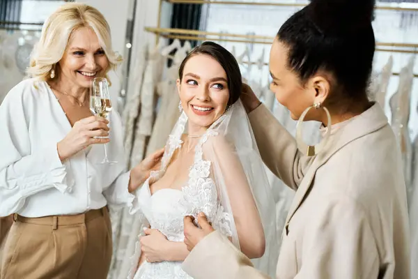 A young brunette bride in a wedding gown is delicately placing a veil on her head, surrounded by her mother and bridesmaid in a bridal salon. — Stock Photo