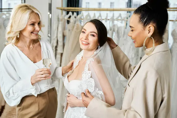 Una joven novia, su madre y la dama de honor de pie con gracia en medio de una variedad de vestidos en un salón de novias. - foto de stock