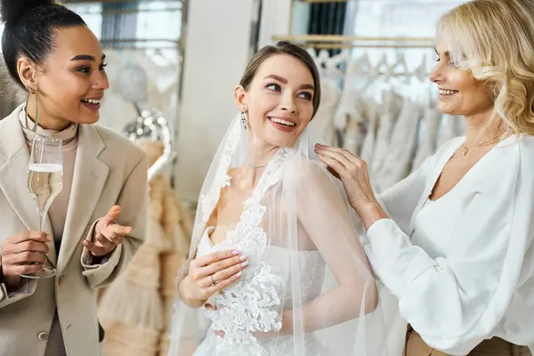 Uma jovem noiva morena em um vestido de casamento excitadamente sorri enquanto sua mãe de meia-idade e melhor amiga, uma dama de honra, ajudá-la a colocar um véu. — Fotografia de Stock