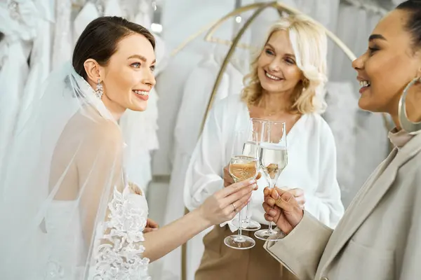 Una joven novia en un vestido de novia y su madre de mediana edad, sosteniendo copas de vino y sonriendo alegremente. - foto de stock