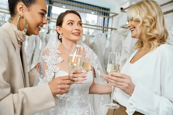 A young brunette bride in a wedding dress, her middle-aged mother, and best friend as a bridesmaid, standing together, holding wine glasses. — Stock Photo