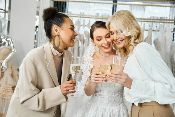 Una joven novia en un vestido de novia, su madre de mediana edad, y la dama de honor se unen, sosteniendo copas de vino. - foto de stock