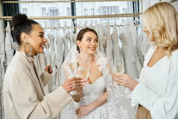 Two brides in wedding attire and a woman with champagne flutes in front of a rack of wedding dresses in bridal salon. — Stock Photo