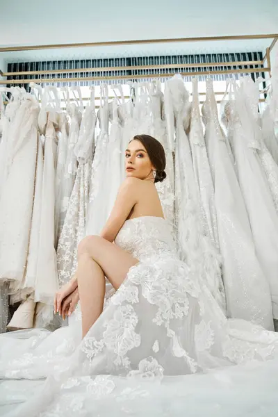 Eine junge brünette Braut sitzt auf einem Bett und starrt auf ein Regal mit Brautkleidern in einem Brautsalon. — Stockfoto