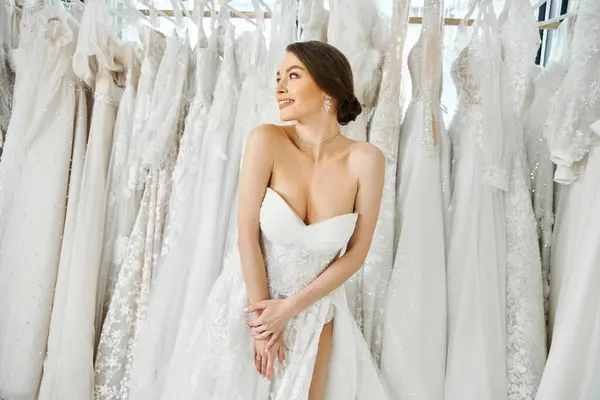 Uma noiva jovem e bonita fica na frente de uma prateleira de vestidos de noiva brancos em um salão de noivas, selecionando cuidadosamente seu vestido.. — Fotografia de Stock