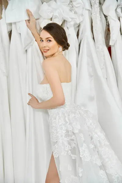 Uma noiva jovem e bonita com cabelo morena fica em um salão de casamento, olhando para um rack de vestidos brancos requintados.. — Fotografia de Stock