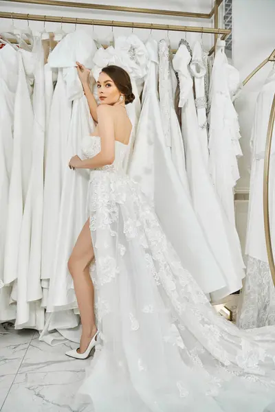Une belle jeune mariée brune debout au milieu d'un rack de robes blanches dans un salon de mariage, à la recherche de la robe parfaite pour sa journée spéciale. — Photo de stock