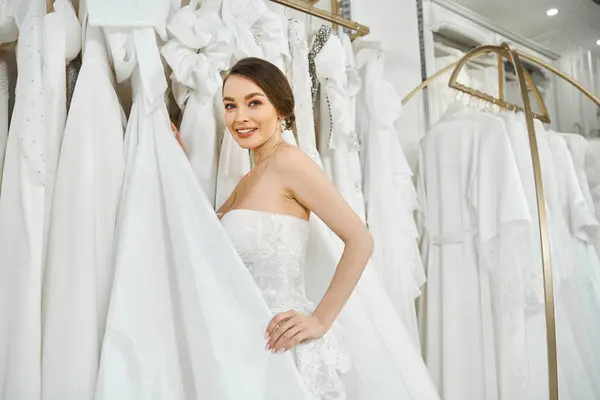 Uma jovem noiva morena fica na frente de uma prateleira de vestidos brancos, escolhendo cuidadosamente seu vestido de casamento perfeito.. — Fotografia de Stock