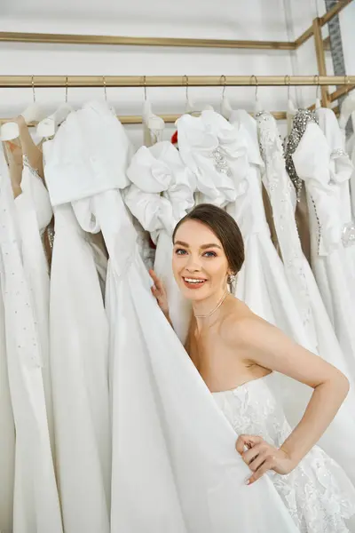Una bella giovane sposa, bruna con un abito bianco fluente, si trova tra una varietà di vestiti in un salone di nozze.. — Foto stock