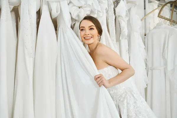 Eine schöne junge Braut, brünett und vor einem Regal mit weißen Kleidern in einem Hochzeitssalon stehend. — Stockfoto
