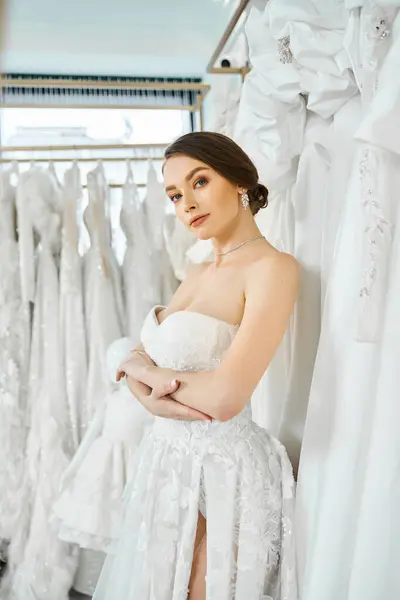 A young brunette bride stands among a rack of dresses in a wedding salon, choosing her perfect gown. — Stock Photo
