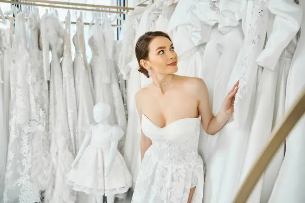 Uma jovem, bela morena de pé na frente de um rack de vestidos em um salão de casamento, escolhendo seu vestido perfeito. — Fotografia de Stock
