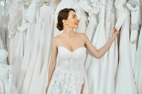 Eine junge, brünette Braut steht zwischen verschiedenen Kleidern in einem Hochzeitssalon. — Stockfoto