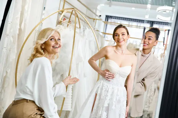 Молода брюнетка в білій весільній сукні стоїть перед дзеркалом поруч з подругою нареченої і матір'ю, приймаючи момент роздумів. — стокове фото