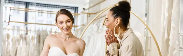 A young brunette bridesmaid and a beautiful bride in a white dress standing next to each other in front of a mirror. — Stock Photo