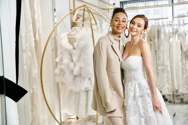 Duas mulheres, uma jovem morena e uma linda noiva em um vestido branco, ficam lado a lado na frente de um rack de vestidos em um salão de casamento.. — Fotografia de Stock