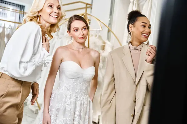 Drei Frauen, eine junge Brünette und eine atemberaubende Braut in einem weißen Kleid stehen gemeinsam vor einem Spiegel. — Stockfoto