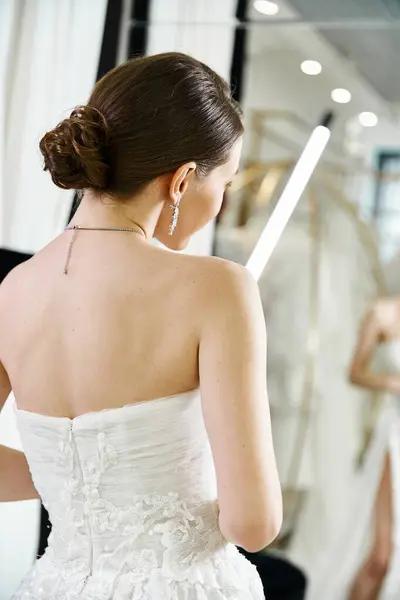 Una joven morena novia en un vestido de novia blanco mira su reflejo en un espejo en un salón de novias. - foto de stock