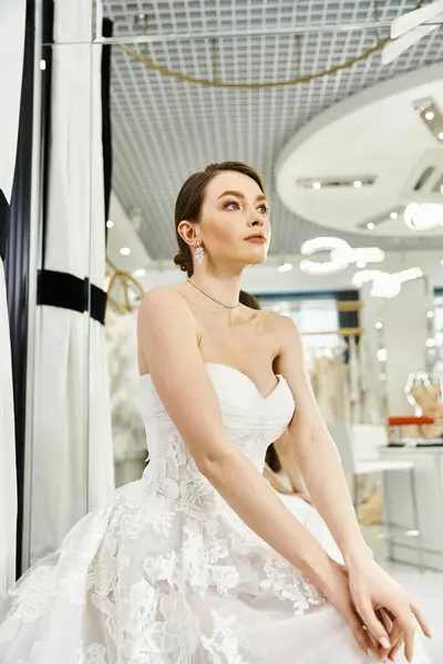 A young, beautiful brunette bride in a flowing white wedding dress sits regally on a chair in a lavish wedding salon. — Stock Photo