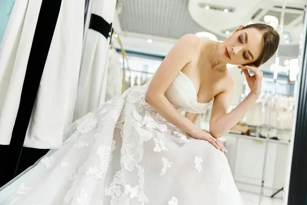 Una joven y hermosa novia morena en un impresionante vestido de novia blanco mira su reflejo en un espejo. - foto de stock