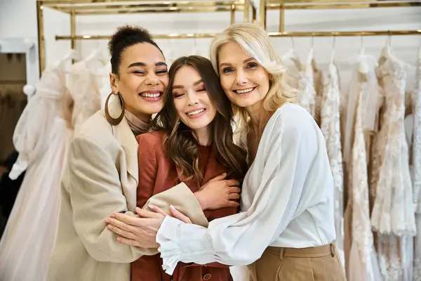Eine junge Braut, ihre Mutter und ihre beste Freundin umarmen sich vor einem Ständer voller Brautkleider, während sie nach dem perfekten Kleid suchen. — Stockfoto