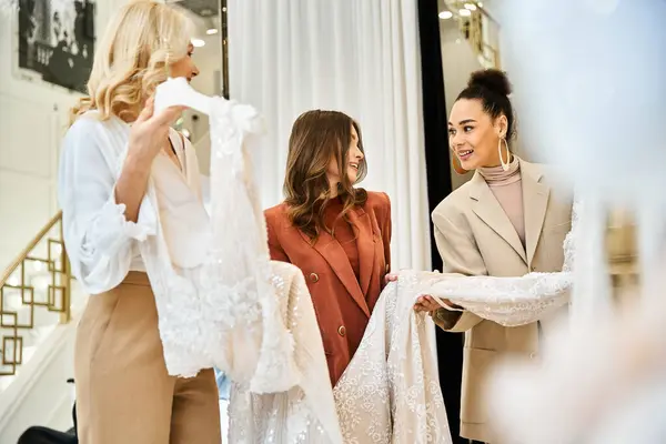 Two women, a mother and best friend, admire a beautiful wedding dress on a mannequin while shopping with a young bride. — Stock Photo