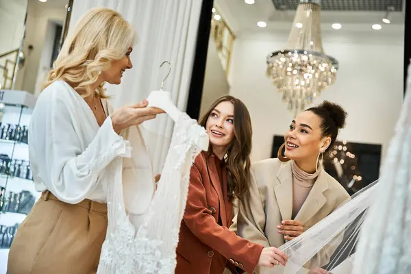 Three women, a young beautiful bride, her mother, and best friend, stand in a store, examining a stunning wedding dress. — Stock Photo