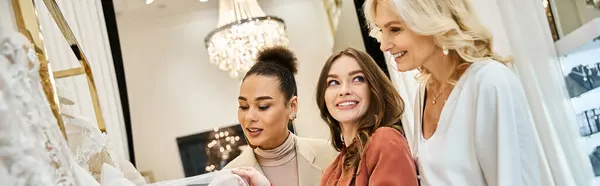 Trois jeunes femmes, dont une belle mariée, examinant une robe de mariée dans un magasin avec concentration et excitation. — Photo de stock