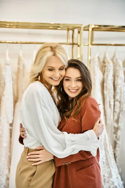 Dos mujeres, una novia y su madre, se abrazan calurosamente en una tienda de vestidos rodeada por un estante de hermosos vestidos. - foto de stock