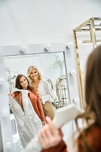 Two women, a young beautiful bride and her mother, carefully selecting wedding dress while looking at themselves in a mirror. — Stock Photo