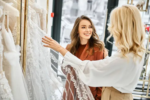 Una joven mujer mira un vestido de novia colgado en un estante, con su madre a su lado, comprando para su próxima boda. - foto de stock