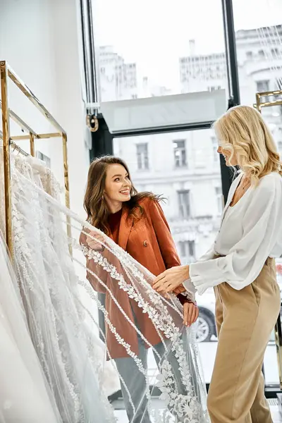 Une jeune belle mariée et sa mère examinent une délicate robe de mariée en dentelle sur un étagère, envisageant le jour spécial des mariées. — Photo de stock