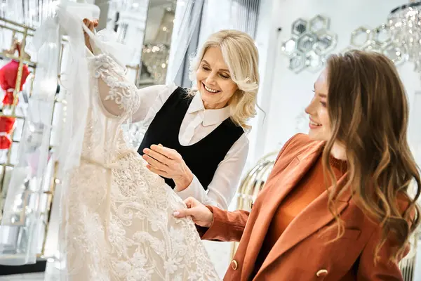 A young beautiful bride and her mother carefully examine a stunning wedding dress in a boutique. — Stock Photo