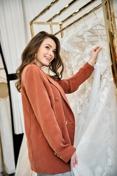 Uma jovem noiva bonita está de pé ao lado de um rack de vestidos, selecionando cuidadosamente seu traje de casamento — Fotografia de Stock