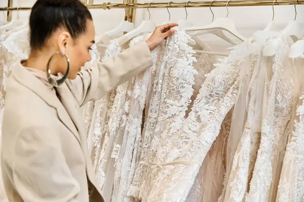 Eine junge, schöne Braut wählt mit Hilfe einer Verkäuferin sorgfältig Brautkleider aus einem vielfältigen Regal aus. — Stockfoto