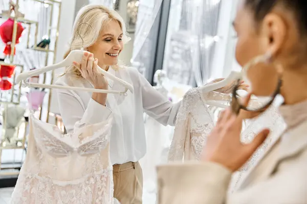 Uma jovem noiva bonita examina cuidadosamente um vestido de noiva em um cabide com a ajuda de um atendente de loja. — Fotografia de Stock