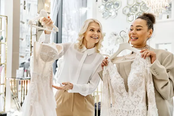 Young women, one a beautiful bride, holding up dresses in a store, assisted by a shop attendant. — Stock Photo