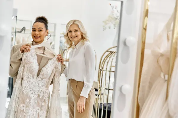 Two women, a young beautiful bride and a shop assistant, stand side by side, admiring themselves in a mirror. — Stock Photo