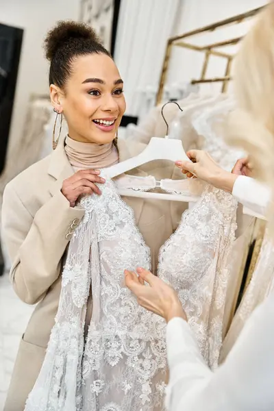 Uma jovem noiva bonita explora um vestido em um cabide enquanto faz compras para seu casamento, assistido por um atendente de loja útil. — Fotografia de Stock